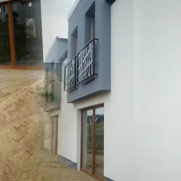 Nowoczesny dom jednorodzinny z białą elewacją, szarymi akcentami i drewnianymi oknami, widok z boku na tle innych domów w trakcie budowy.