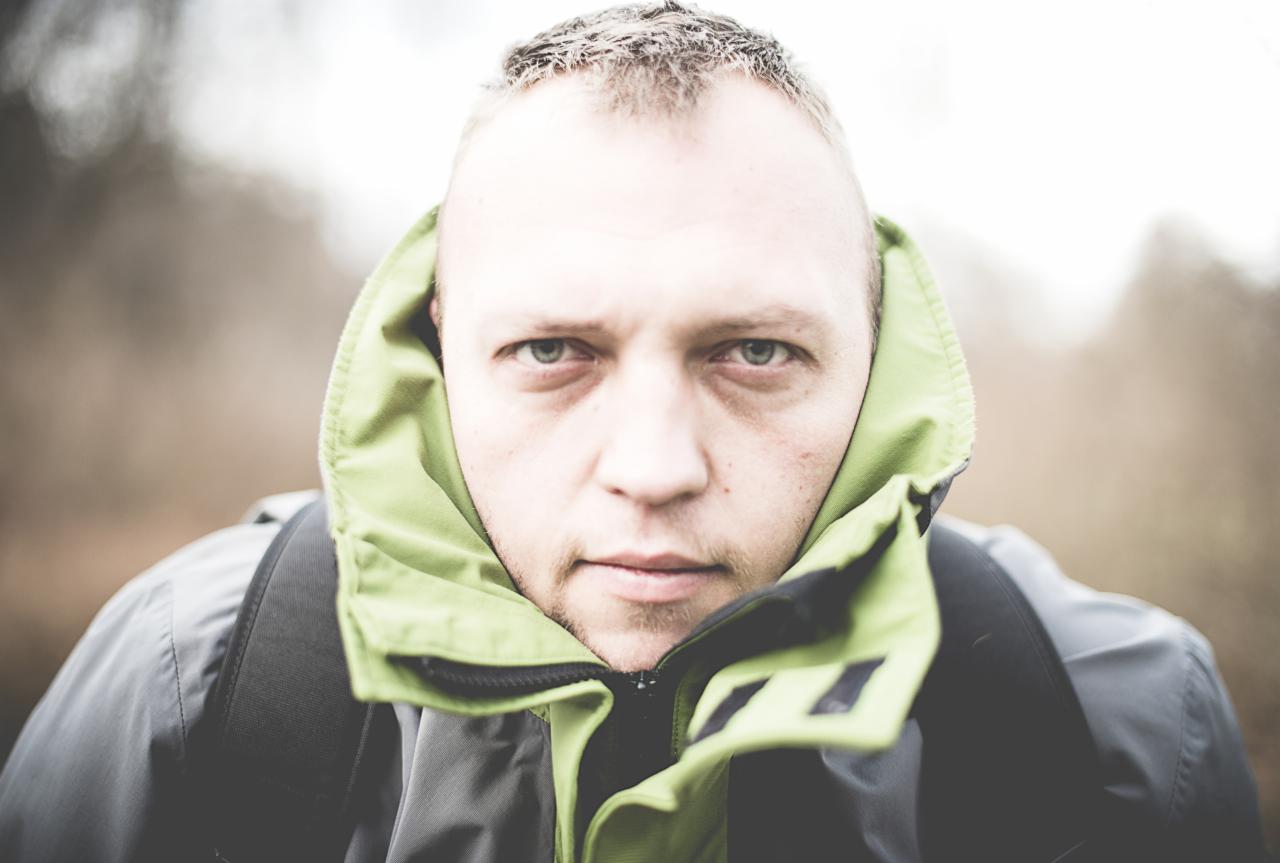 Portret mężczyzny w szaro-zielonej kurtce z kapturem, patrzącego wprost w obiektyw, z plecakiem na ramionach, na rozmytym tle.