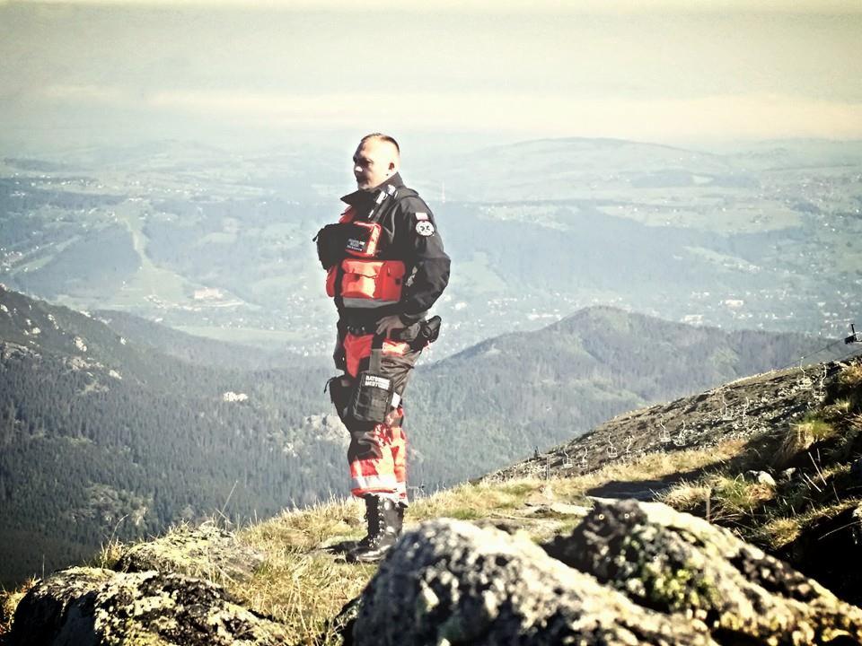 Ratownik górski w uniformie i z apteczką, stoi na szczycie góry z widokiem na dolinę.