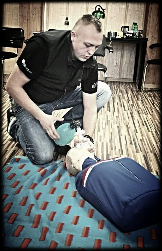 Instruktor w czarnej kamizelce i jeansach klęczy na podłodze z drewnianymi panelami, demonstrując resuscytację na manekinie ułożonym na turkusowym kocu w ceglaste wzory, używając worka...