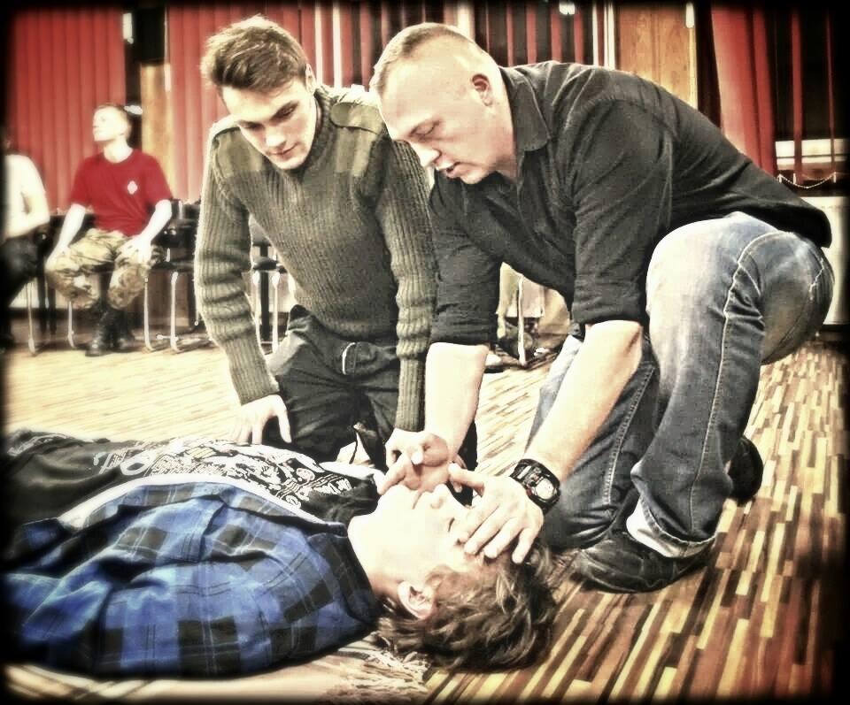 Instruktorzy prowadzą resuscytację krążeniowo-oddechową na manekinie podczas szkolenia w Zabrzu, z widocznym zegarkiem na ręce jednego z nich i obserwującymi uczestnikami w tle.