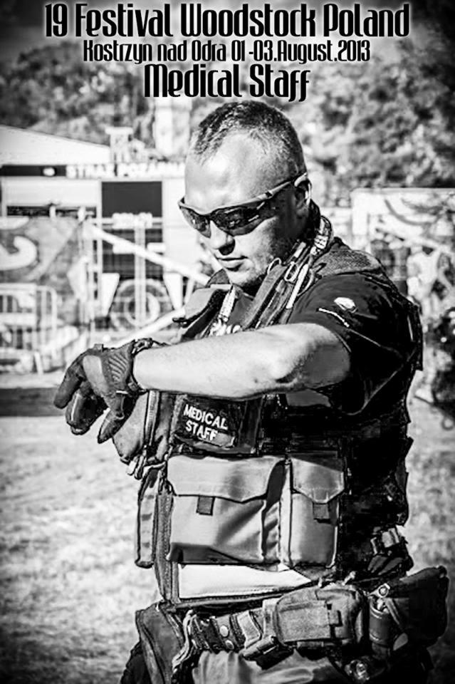Czarno-białe zdjęcie ratownika medycznego w kamizelce taktycznej z napisem 'MEDICAL STAFF' sprawdzającego godzinę na zegarku podczas 19. Festiwalu Woodstock Poland w Kostrzynie nad Odrą w sierpniu...