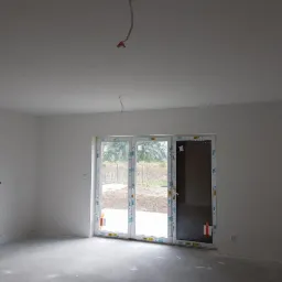 Wnętrze w stanie deweloperskim z gładkimi, białymi ścianami i oknami balkonowymi wychodzącymi na ogród.