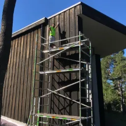 Mężczyzna na rusztowaniu maluje drewnianą elewację budynku na ciemny kolor, widoczne narzędzia i częściowo pomalowana ściana, w tle las i błękitne niebo.