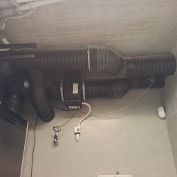 Recohouse - Czarna, elastyczna instalacja wentylacyjna z centralą Vasco DX6, zamontowana pod betonowym sufitem w pomieszczeniu z szarą, ceramiczną ścianą.