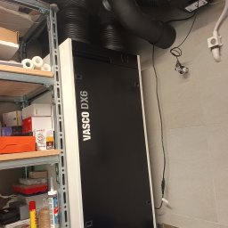 Recohouse - Czarna jednostka rekuperacyjna Vasco DX6 z podłączonymi przewodami wentylacyjnymi, zainstalowana w pomieszczeniu w Pruszkowie. Widoczne rury i elementy montażowe.