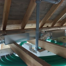 Recohouse - Zielone rury wentylacyjne pod drewnianym stropem, srebrne kanały izolowane folią aluminiową i szara rura pionowa. Instalacja rekuperacji w trakcie montażu.