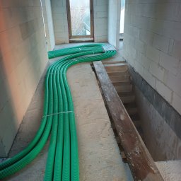 Recohouse - Zielone rury wentylacyjne ułożone w surowym wnętrzu budynku z białych bloczków, widoczne schody i deska. Instalacja rekuperacji w trakcie montażu.