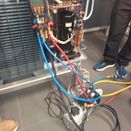 Recohouse - Podłączenie manometrów do instalacji chłodniczej. Widoczne przewody, miernik i elementy instalacji. Prace serwisowe w toku, z fragmentami sylwetek osób w tle.