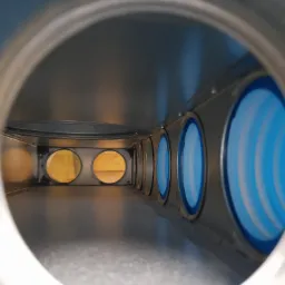 Wnętrze metalowego kanału wentylacyjnego z okrągłymi otworami, z których niektóre są wypełnione żółtym materiałem izolacyjnym, widziane z perspektywy wnętrza kanału.