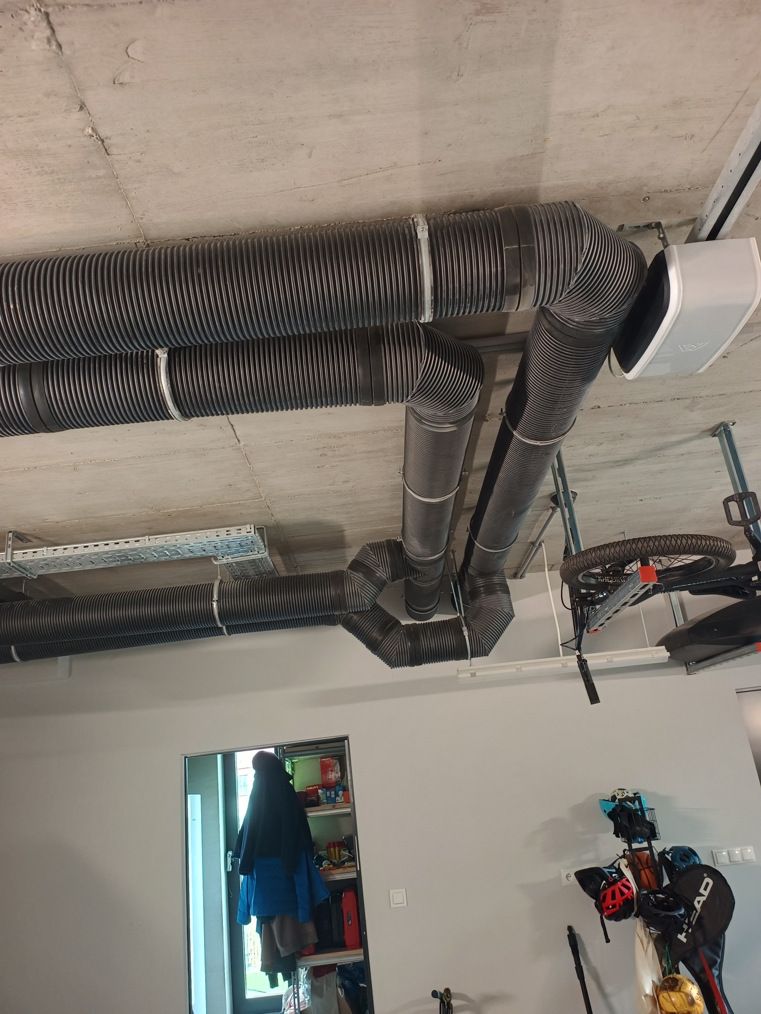 Szare, elastyczne rury wentylacyjne pod betonowym sufitem, zamontowane w garażu. Widoczny biały moduł rekuperatora i fragment roweru podwieszonego pod sufitem.