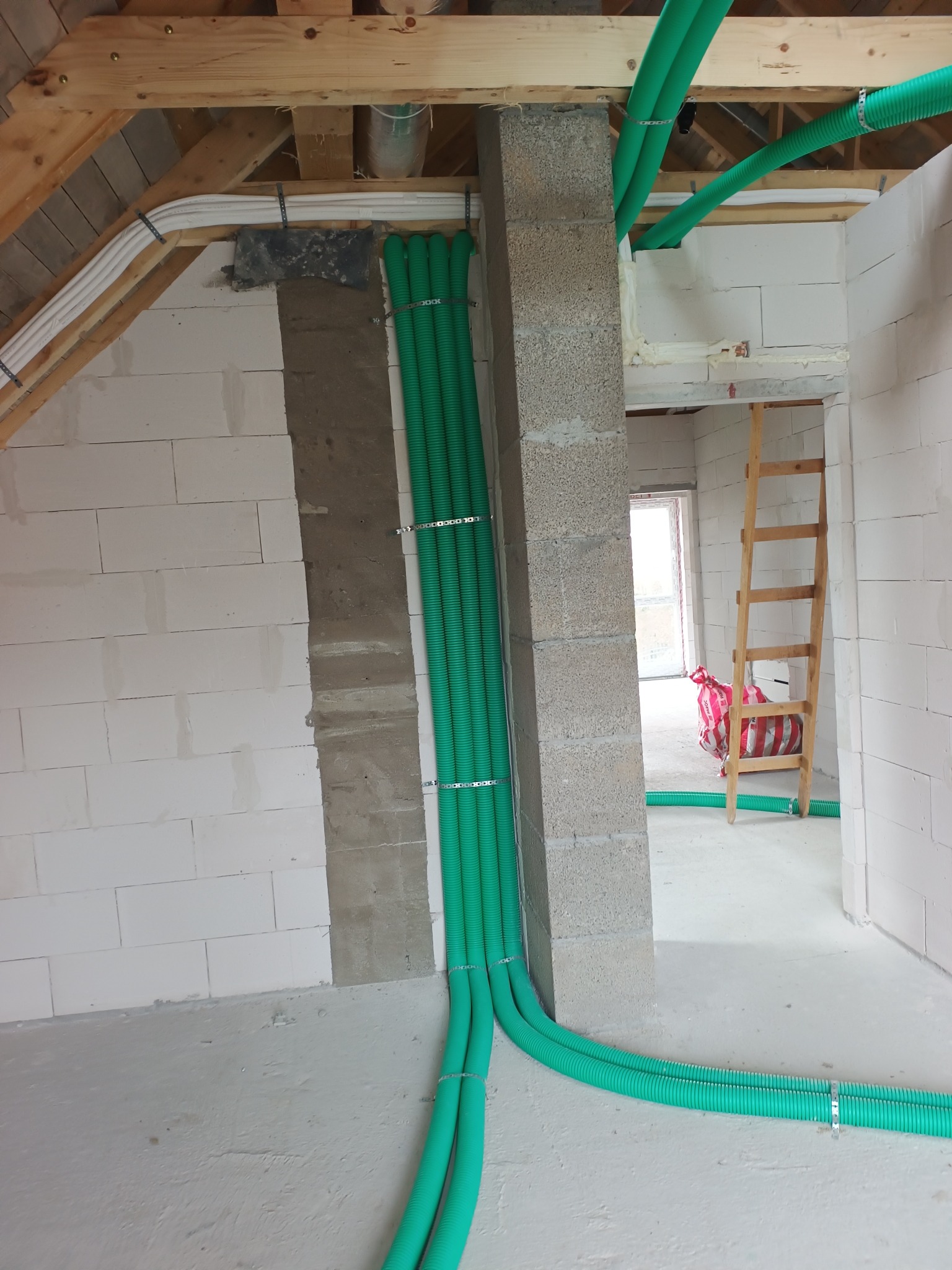 Zielone rury wentylacyjne przymocowane do ściany z betonu komórkowego w surowym wnętrzu budynku w trakcie budowy. Widoczne drewniane elementy konstrukcji dachu.