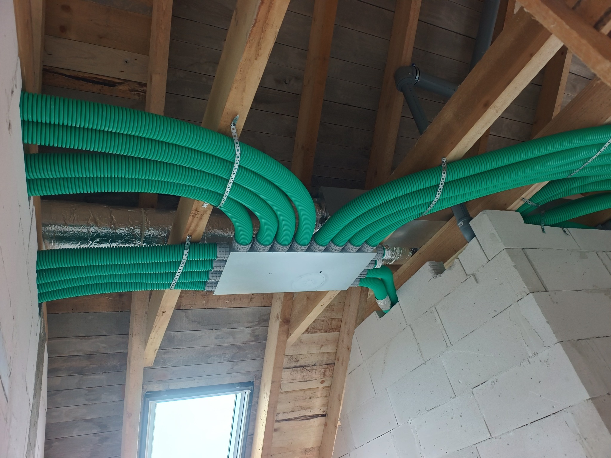 Zielone, karbowane rury wentylacyjne zamontowane pod drewnianym stropem, widoczne elementy instalacji i białe bloczki. Instalacja w trakcie montażu.