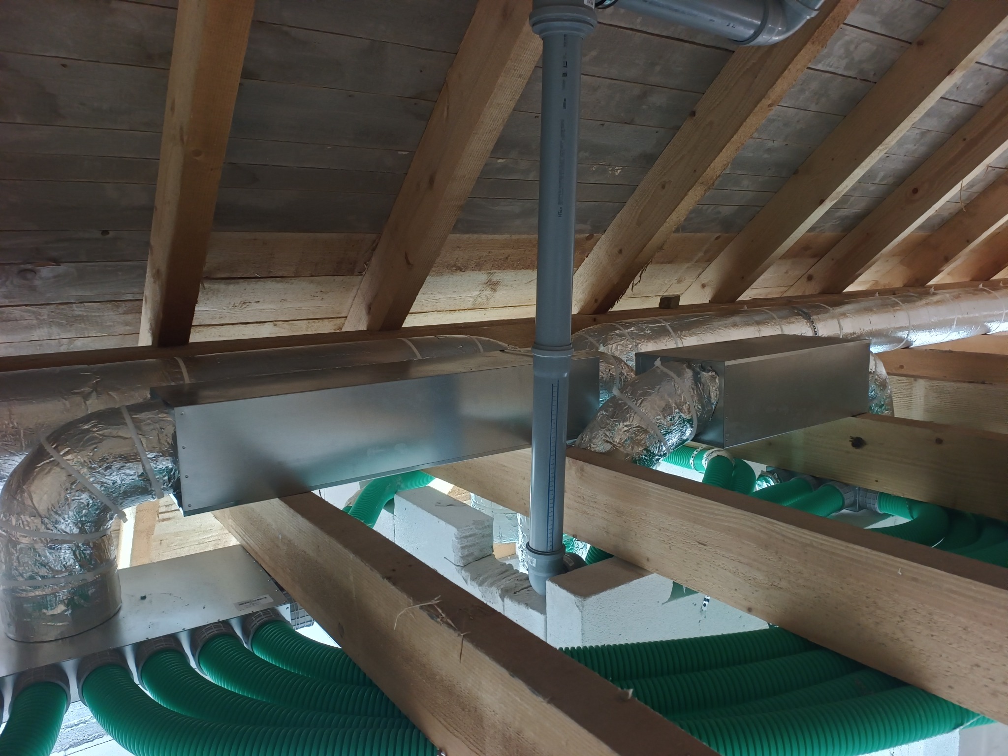 Zielone rury wentylacyjne pod drewnianym stropem, srebrne kanały izolowane folią aluminiową i szara rura pionowa. Instalacja rekuperacji w trakcie montażu.