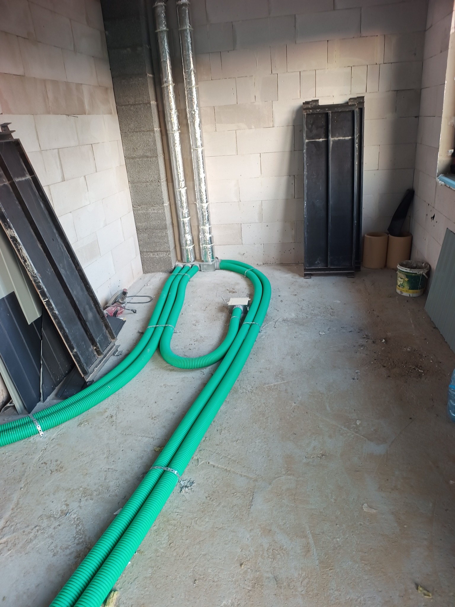 Wnętrze w trakcie montażu wentylacji: widoczne zielone rury wentylacyjne na podłodze, srebrne rury pionowe przy ścianie z betonu komórkowego i elementy instalacyjne.