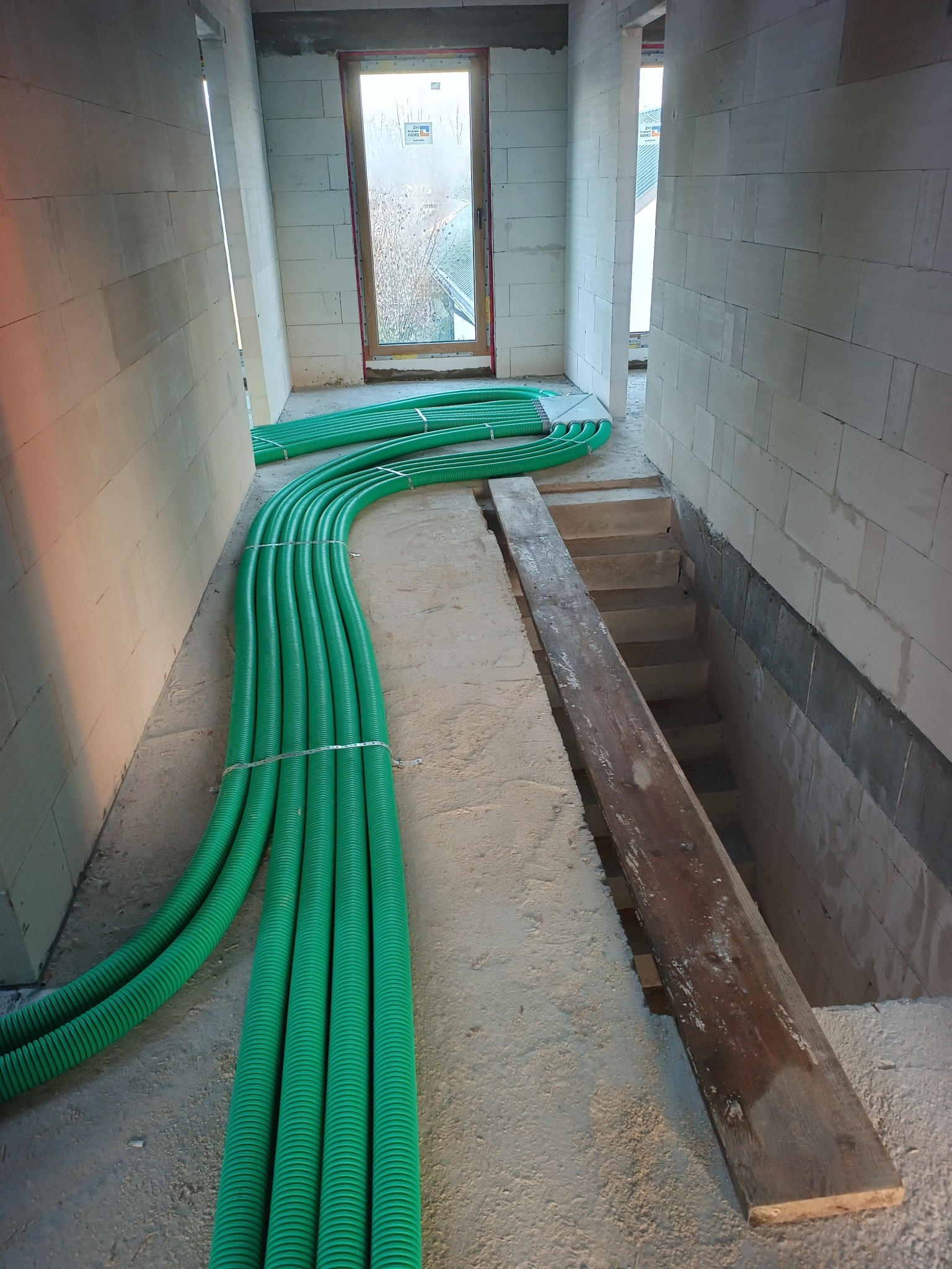 Zielone rury wentylacyjne ułożone w surowym wnętrzu budynku z białych bloczków, widoczne schody i deska. Instalacja rekuperacji w trakcie montażu.