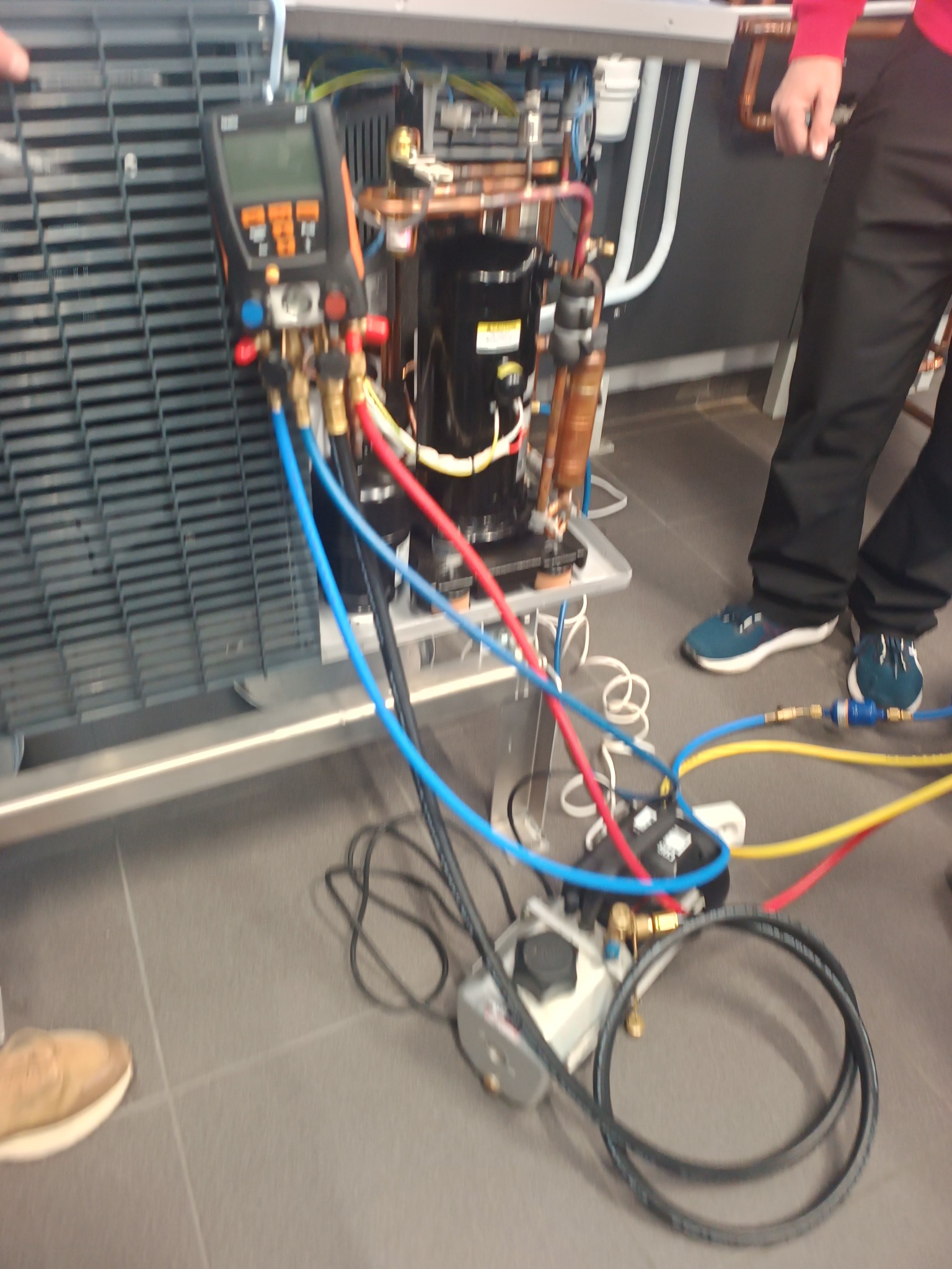 Podłączenie manometrów do instalacji chłodniczej. Widoczne przewody, miernik i elementy instalacji. Prace serwisowe w toku, z fragmentami sylwetek osób w tle.