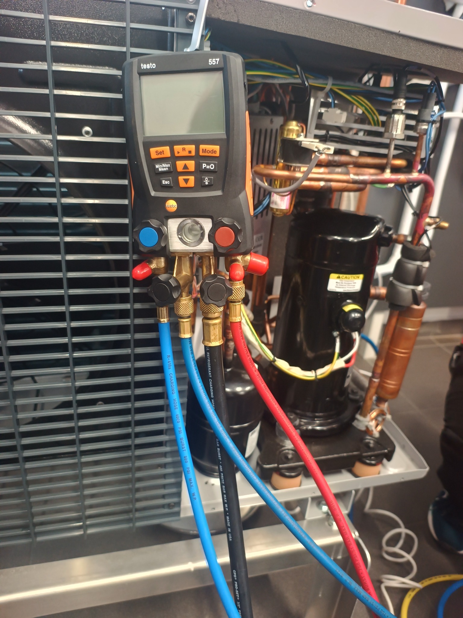 Miernik Testo 557 podłączony do instalacji chłodniczej z widocznym kompresorem i przewodami. Narzędzie diagnostyczne w trakcie pomiarów parametrów pracy układu.