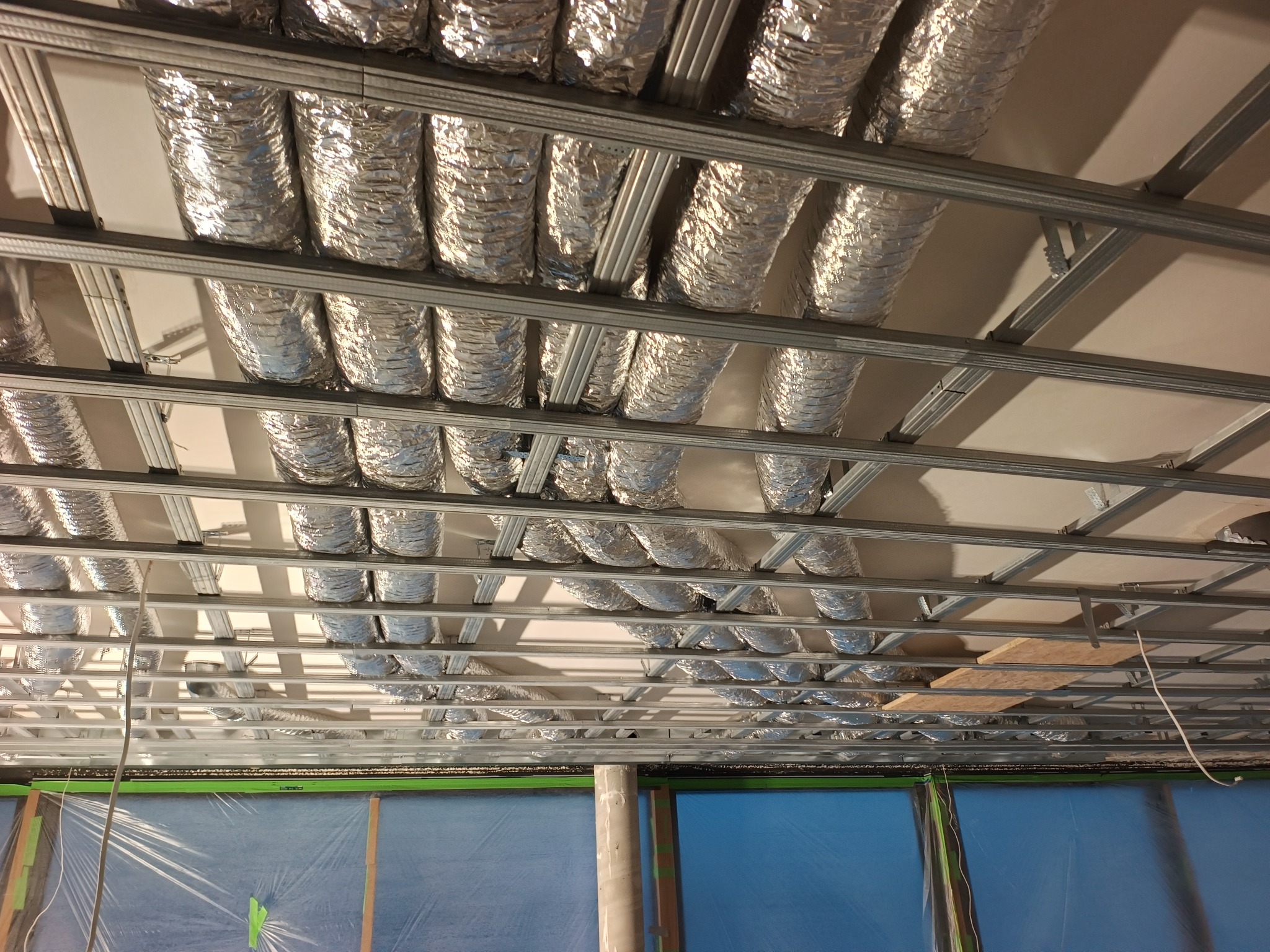 Srebrne, elastyczne przewody wentylacyjne zamontowane pod sufitem podwieszanym z metalową konstrukcją. Ściany zabezpieczone folią. Widok z dołu.