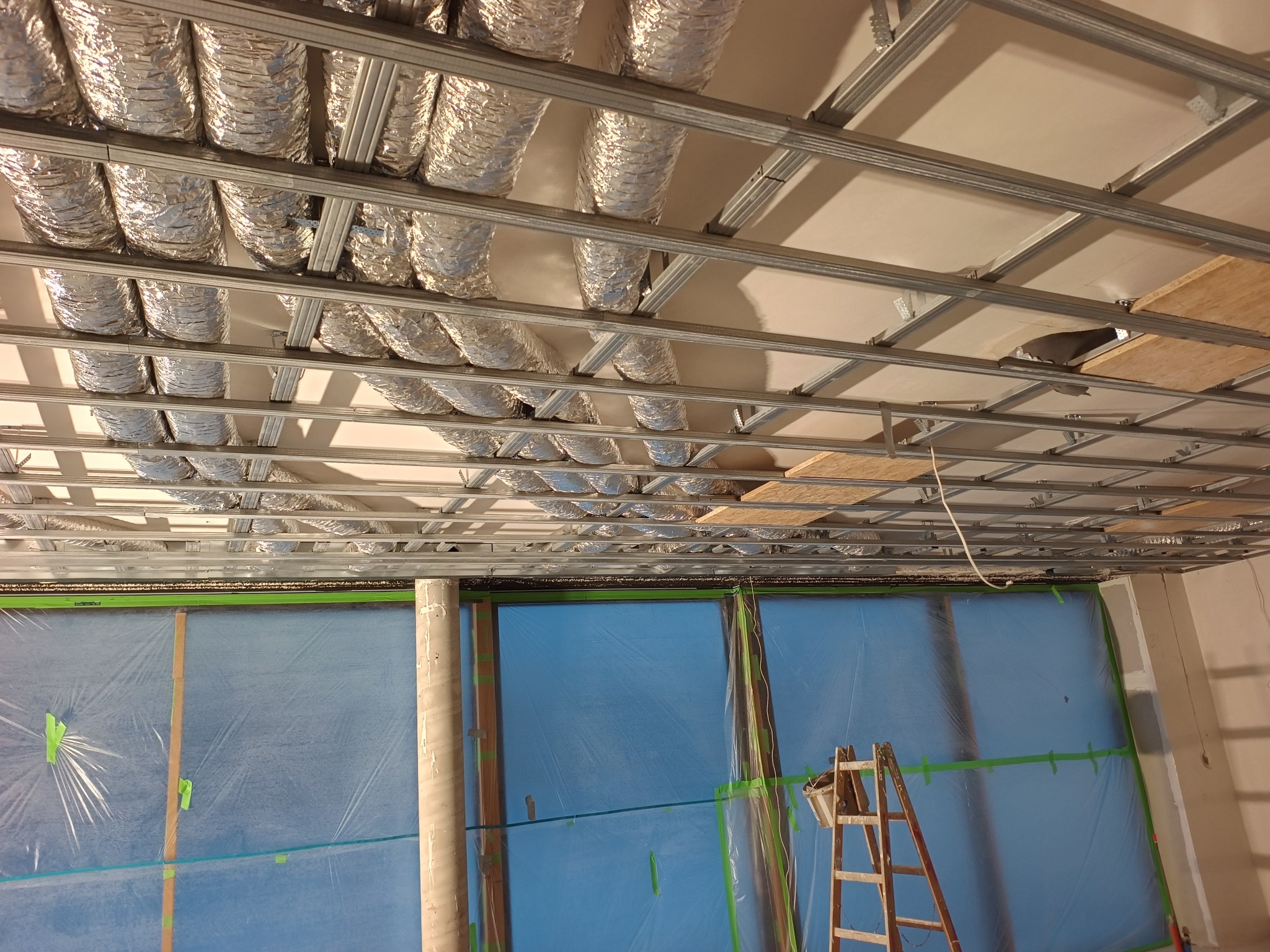 Montaż instalacji wentylacyjnej: srebrne rury pod sufitem, metalowa konstrukcja, zabezpieczone folią okna, drabina. Widok z dołu, etap prac.