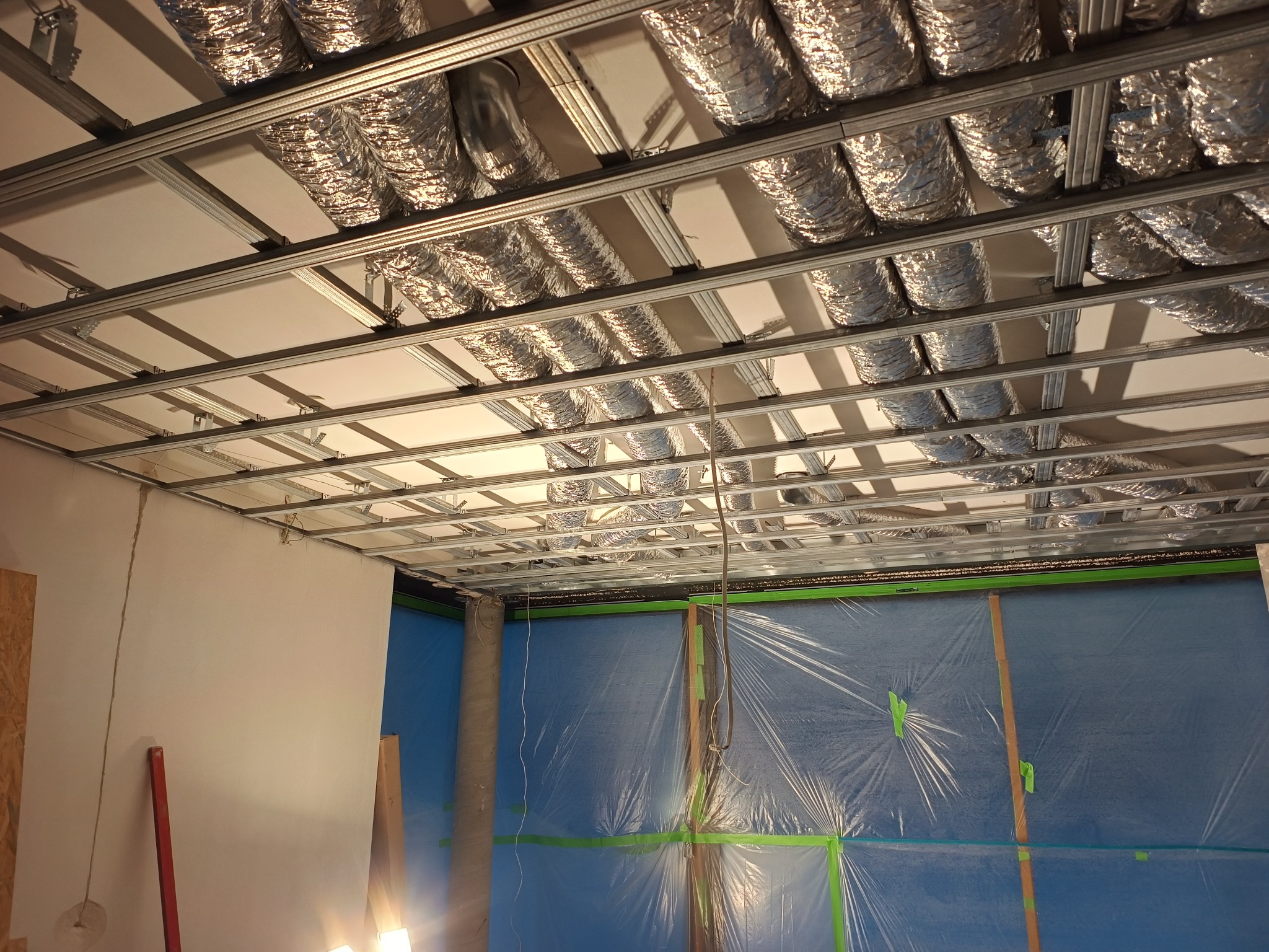 Srebrne rury wentylacyjne pod sufitem z metalową konstrukcją. Ściany zabezpieczone folią. Widok instalacji w trakcie montażu.