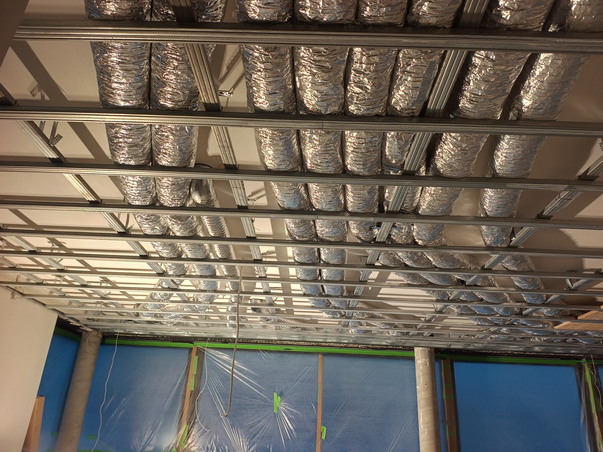 Srebrne, elastyczne przewody wentylacyjne zamontowane pod sufitem podwieszanym, widoczne elementy konstrukcji i zabezpieczona folią ściana. Instalacja w trakcie prac.