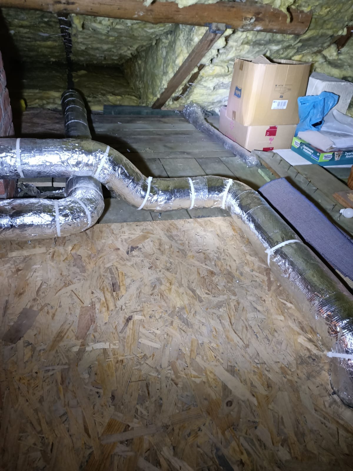 Instalacja srebrnych, elastycznych przewodów wentylacyjnych w przestrzeni poddasza, zabezpieczonych białymi opaskami zaciskowymi. Widoczne drewniane elementy konstrukcyjne.