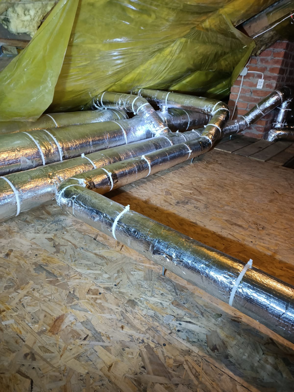 Izolowane rury wentylacyjne z licznymi złączami, zamocowane na poddaszu, widoczne elementy konstrukcji dachu i ściana z cegły. Instalacja w trakcie montażu.