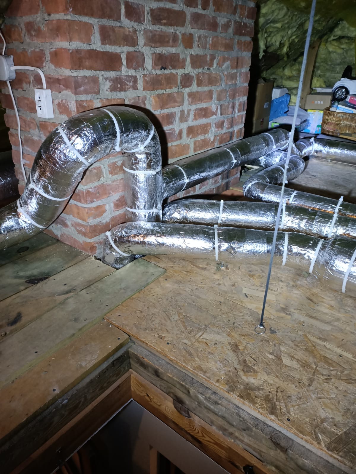 Srebrne, izolowane rury wentylacyjne przymocowane białymi opaskami, ułożone na drewnianej podłodze poddasza, z fragmentem ceglanej ściany w tle. Instalacja w trakcie montażu.