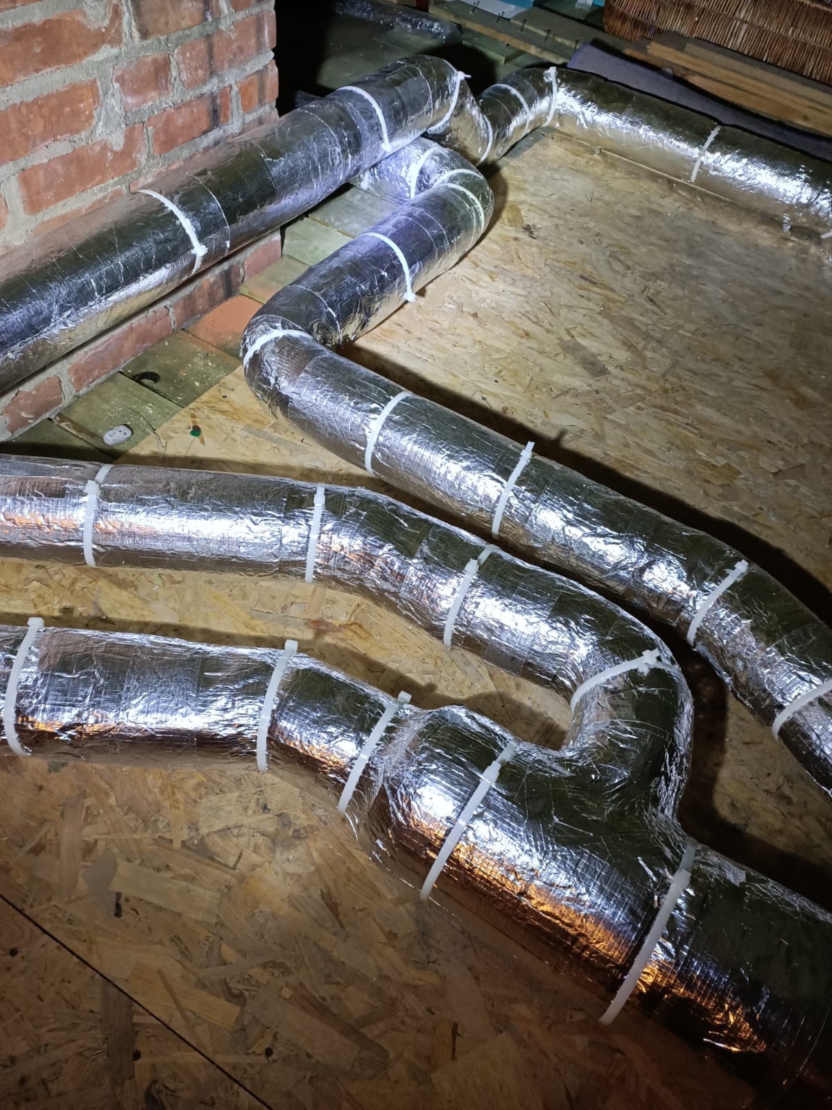 Izolowane, srebrne rury wentylacyjne z białymi opaskami zaciskowymi, ułożone na drewnianej powierzchni, z fragmentem ceglanej ściany w tle. Instalacja w trakcie montażu.