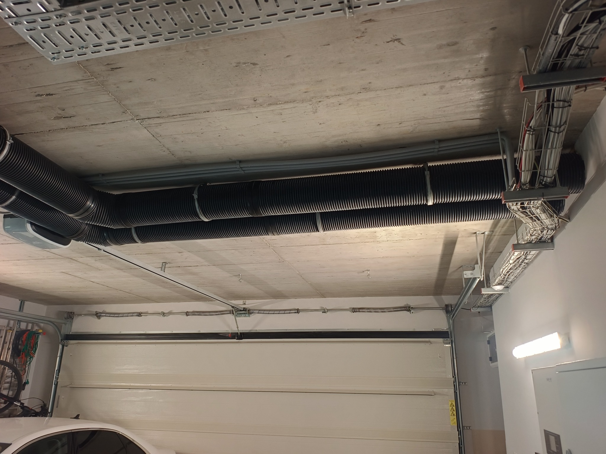 Czarna, karbowana instalacja wentylacyjna pod betonowym sufitem garażu, z widocznymi elementami mocującymi i przewodami elektrycznymi. W tle brama garażowa i fragment samochodu.