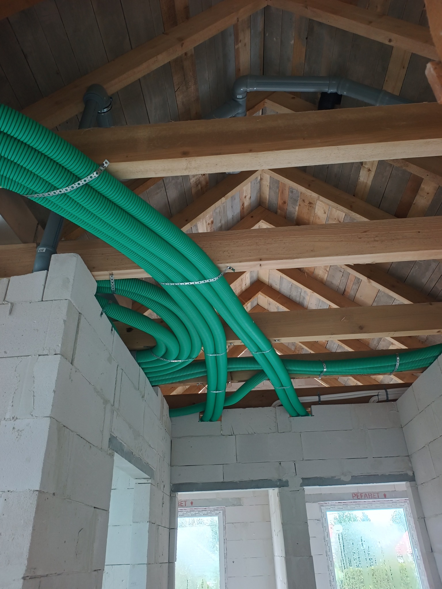 Zielone rury wentylacyjne zamontowane pod drewnianym stropem w surowym wnętrzu budynku z białych bloczków. Widoczne elementy mocujące i fragmenty instalacji.