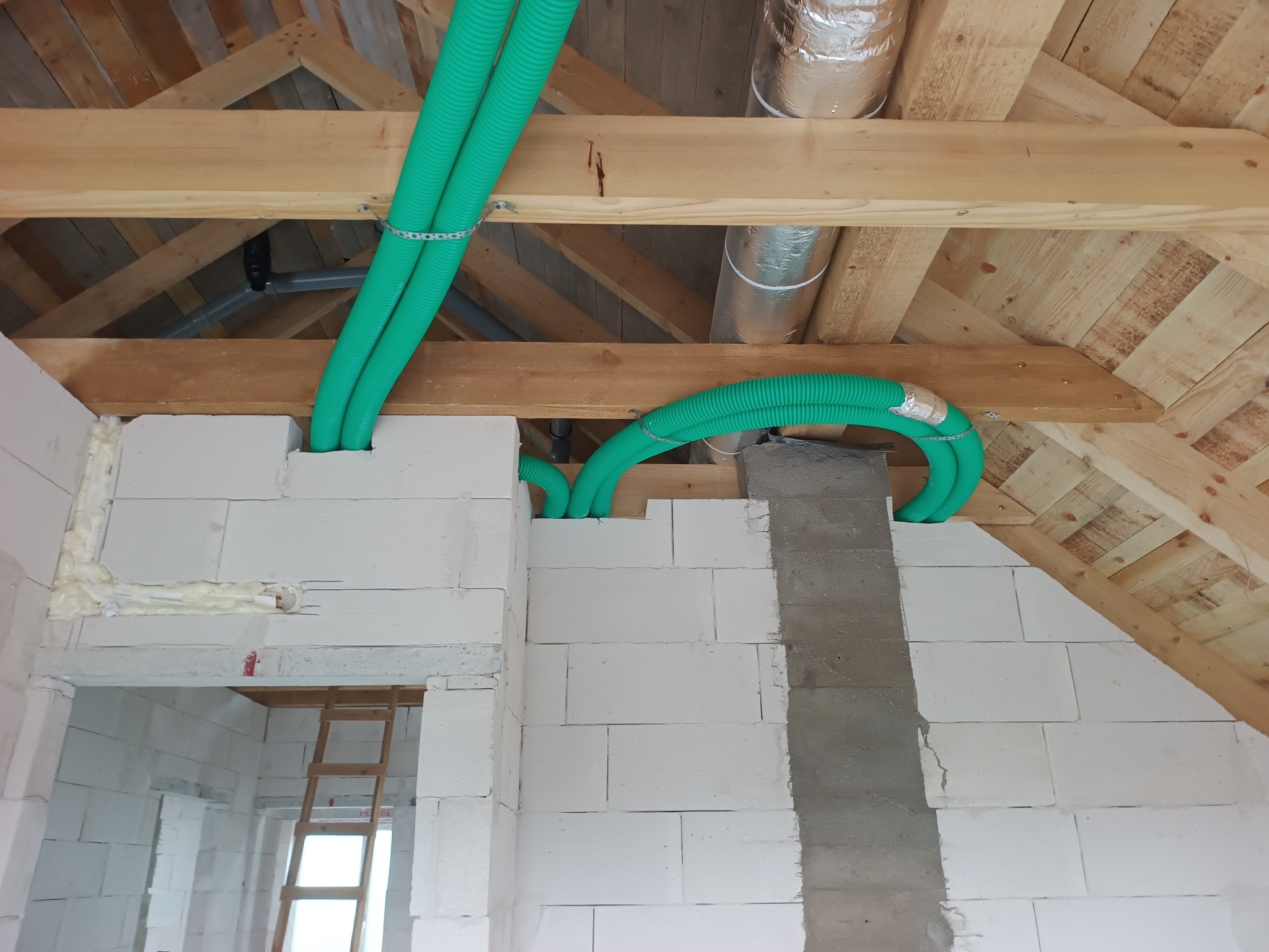 Zielone rury wentylacyjne zamontowane pod drewnianym stropem na poddaszu budynku w trakcie budowy. Widoczne białe bloczki i fragment komina.