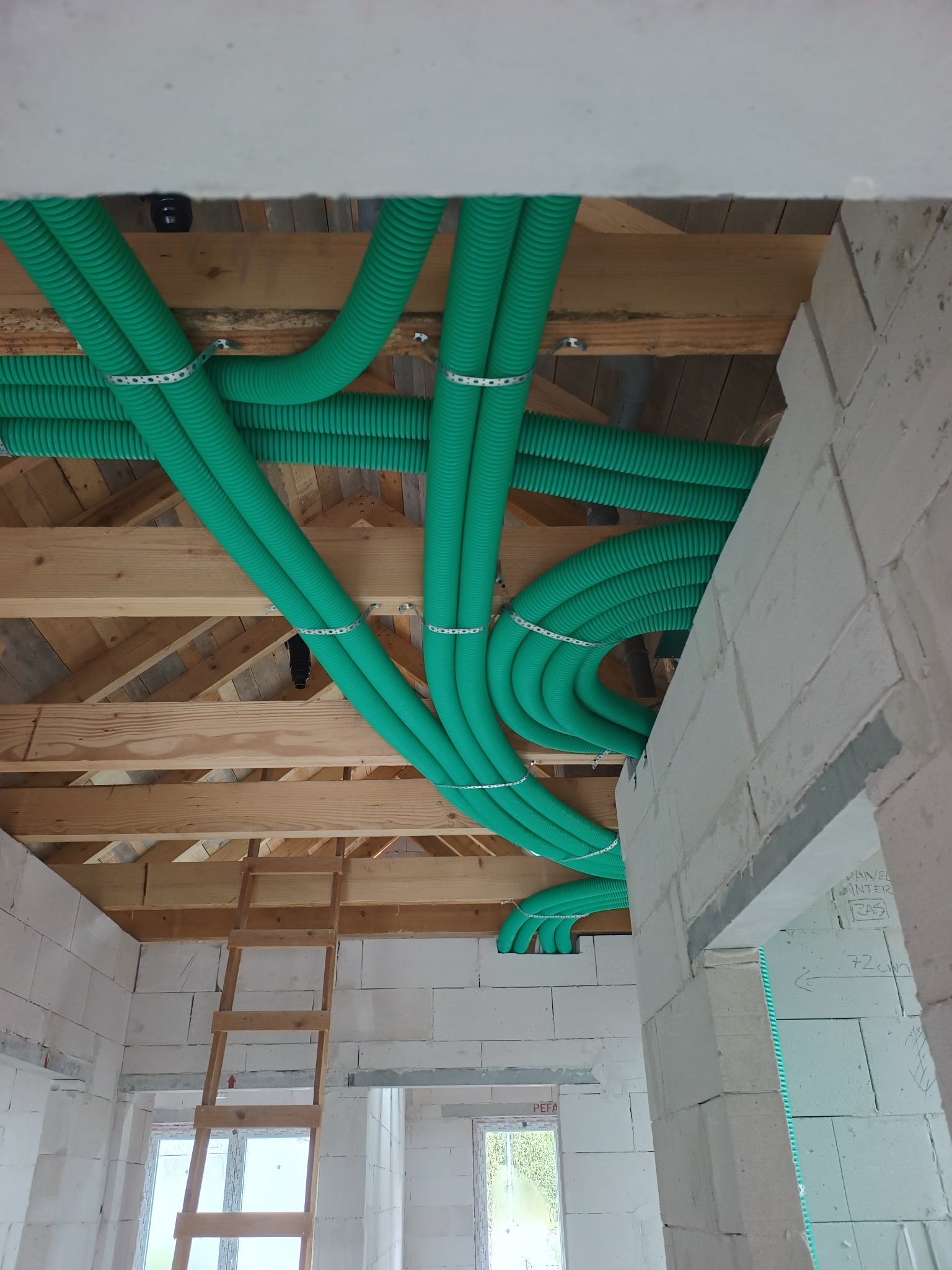 Zielone rury wentylacyjne zamontowane pod drewnianym stropem w surowym wnętrzu budynku z białych bloczków, widoczne mocowania i drabina.