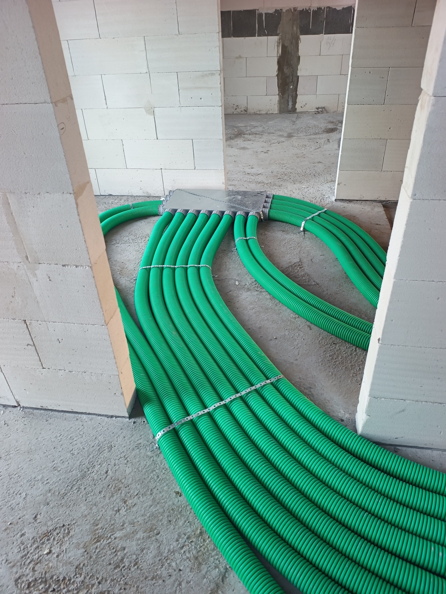 Zielone rury wentylacyjne ułożone równolegle na surowym betonie, widoczne między ścianami z białych bloczków. Instalacja w trakcie budowy, surowy stan.