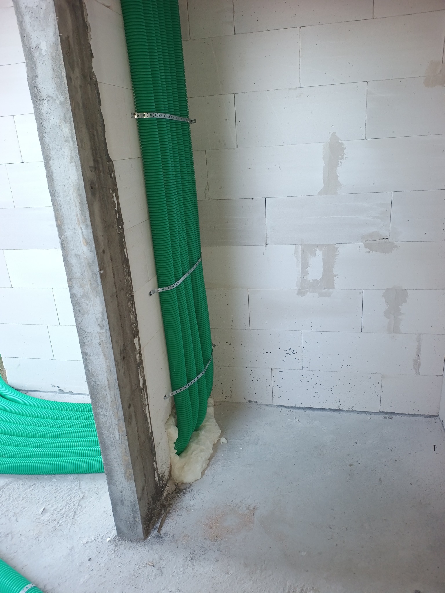 Zielone rury wentylacyjne przymocowane do ściany z betonu komórkowego, zabezpieczone pianką montażową. Widoczne metalowe uchwyty mocujące. Surowy stan wykończenia wnętrza.