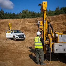 Pracownik w kasku obsługuje żółtą wiertnicę Staltechnika na tle wykopu i białego pickupa CGI z otwartymi drzwiami. Błękitne niebo.