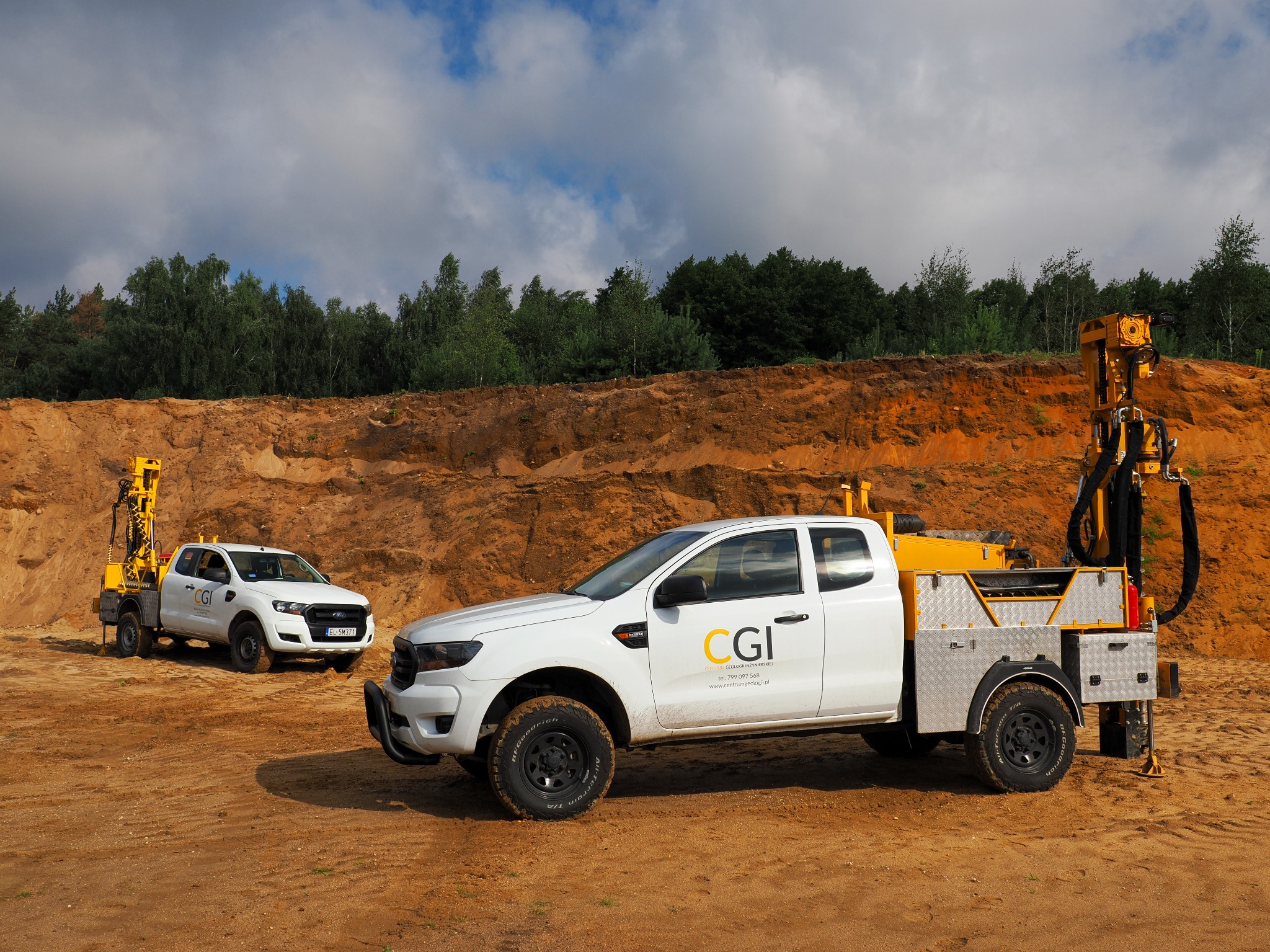 Dwa białe samochody terenowe z wiertnicami na piaszczystym terenie. Na pierwszym planie Ford Ranger z logo firmy CGI. W tle wykop i drzewa.