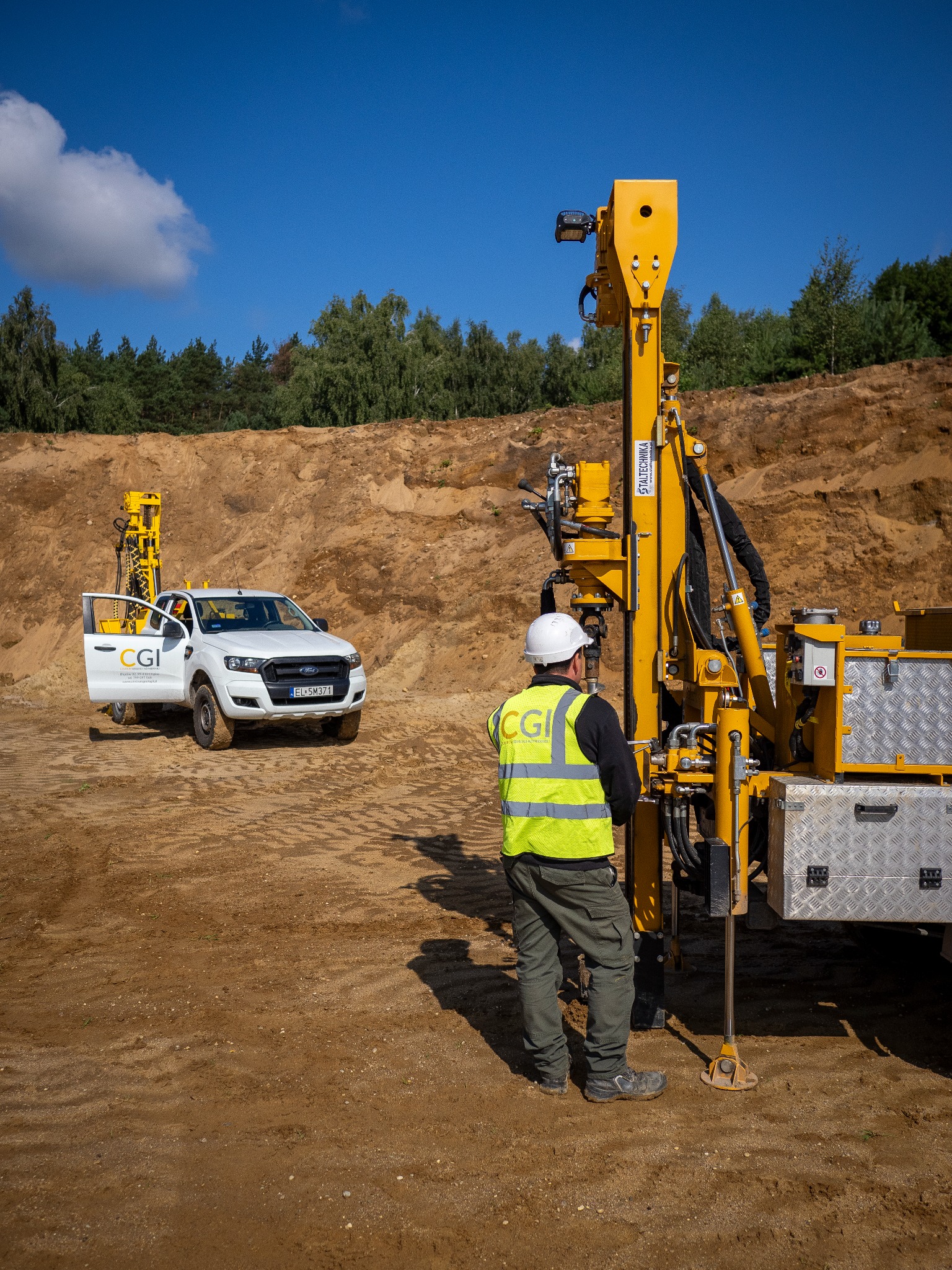 Pracownik w kasku obsługuje żółtą wiertnicę Staltechnika na tle wykopu i białego pickupa CGI z otwartymi drzwiami. Błękitne niebo.