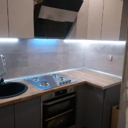 Nowoczesna, szara kuchnia z drewnianym blatem, płytą gazową i czarnym zlewem, oświetlona listwą LED pod szafkami wiszącymi i z widocznym fragmentem niezabezpieczonej ściany w prawym dolnym rogu.