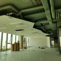 Montaż sufitu podwieszanego w biurze: widoczne płyty sufitowe, rury wentylacyjne, drabina i okna z widokiem na miasto. Etap prac wykończeniowych.