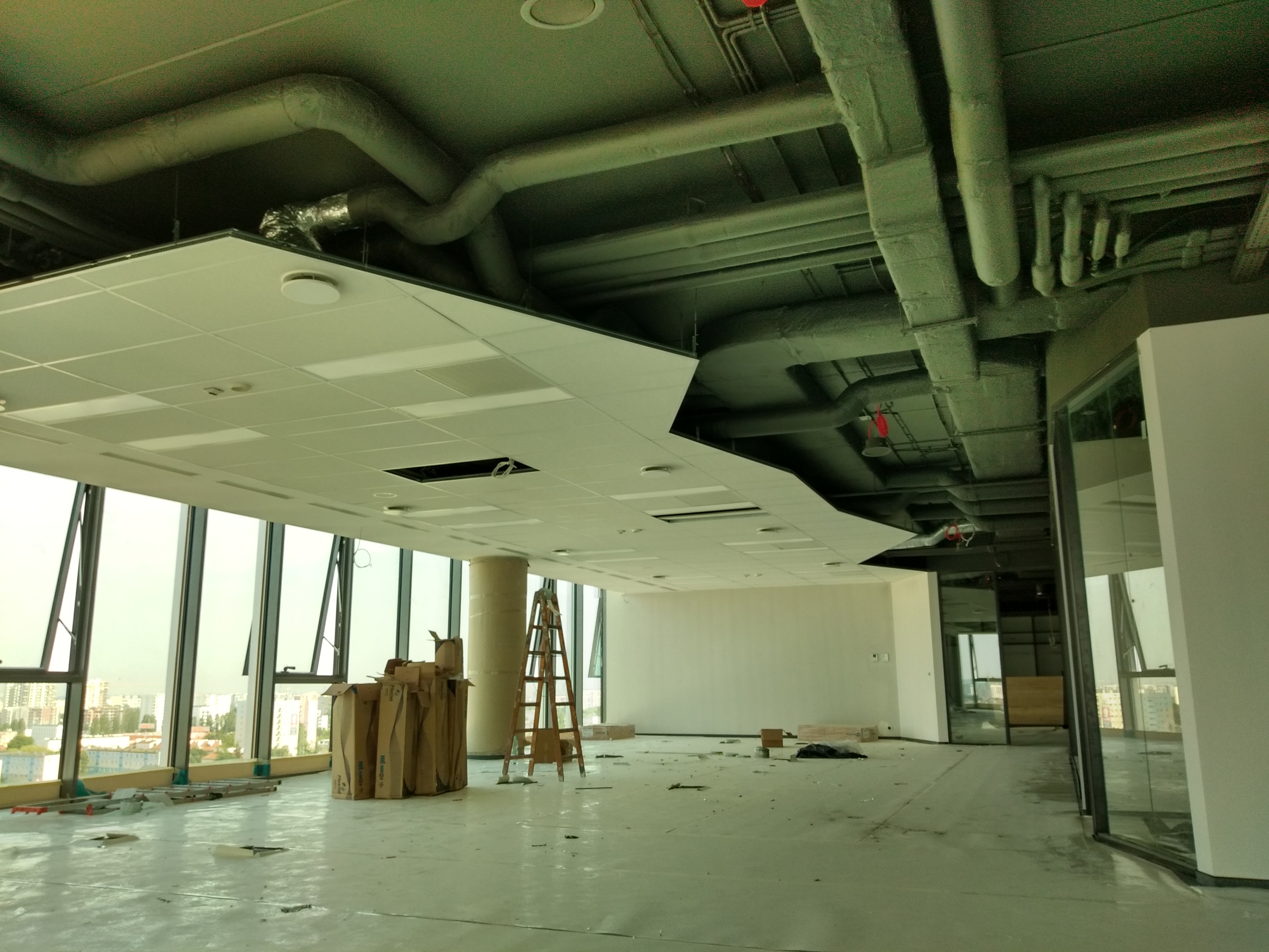 Montaż sufitu podwieszanego w biurze: widoczne płyty sufitowe, rury wentylacyjne, drabina i okna z widokiem na miasto. Etap prac wykończeniowych.