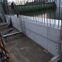 Betonowy mur oporowy z wystającymi prętami zbrojeniowymi, widoczny z góry, z fragmentem rzeki w tle i pracownikiem w pomarańczowej kurtce na górze konstrukcji.