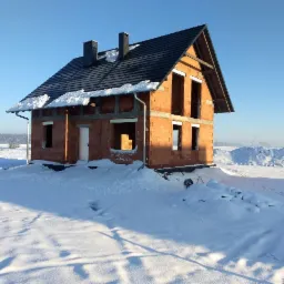 Surowy, dwukondygnacyjny dom z czerwonej cegły w trakcie budowy, z dachem pokrytym ciemną dachówką, otoczony śnieżnym krajobrazem w słoneczny dzień.