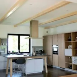 Jasna kuchnia z drewnianymi belkami stropowymi, wyspą z blatem roboczym i szafkami w odcieniach beżu, widziana z perspektywy podłogi.