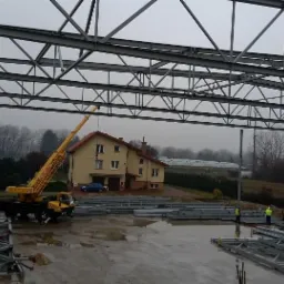 Stalowa konstrukcja hali w trakcie montażu z użyciem dźwigu, widok na budynek mieszkalny w tle. Elementy konstrukcyjne na mokrym placu budowy.