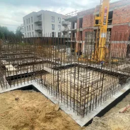 Zbrojenie fundamentów pod budynek wielorodzinny, widoczny szalunek i stalowe pręty, w tle budynek w stanie surowym i dźwig budowlany.