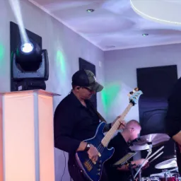 Muzycy grający na instrumentach podczas występu: basista w czarnej koszuli i czapce z daszkiem gra na niebieskiej gitarze basowej, wokalista śpiewa do mikrofonu, a perkusista gra na perkusji.