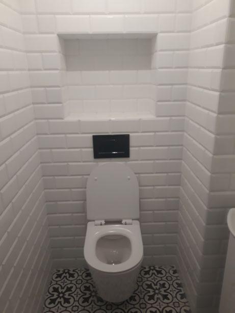 Toaleta z otwartą klapą w małej łazience wykończonej białymi, prostokątnymi płytkami imitującymi cegłę i wzorzystą posadzką.