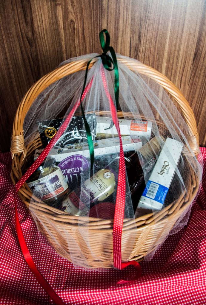 Wiklinowy kosz wypełniony różnorodnymi produktami spożywczymi, takimi jak słodycze, konfitury i napoje, owinięty tiulem i ozdobiony czerwonymi i zielonymi wstążkami, ustawiony na czerwono-białej...